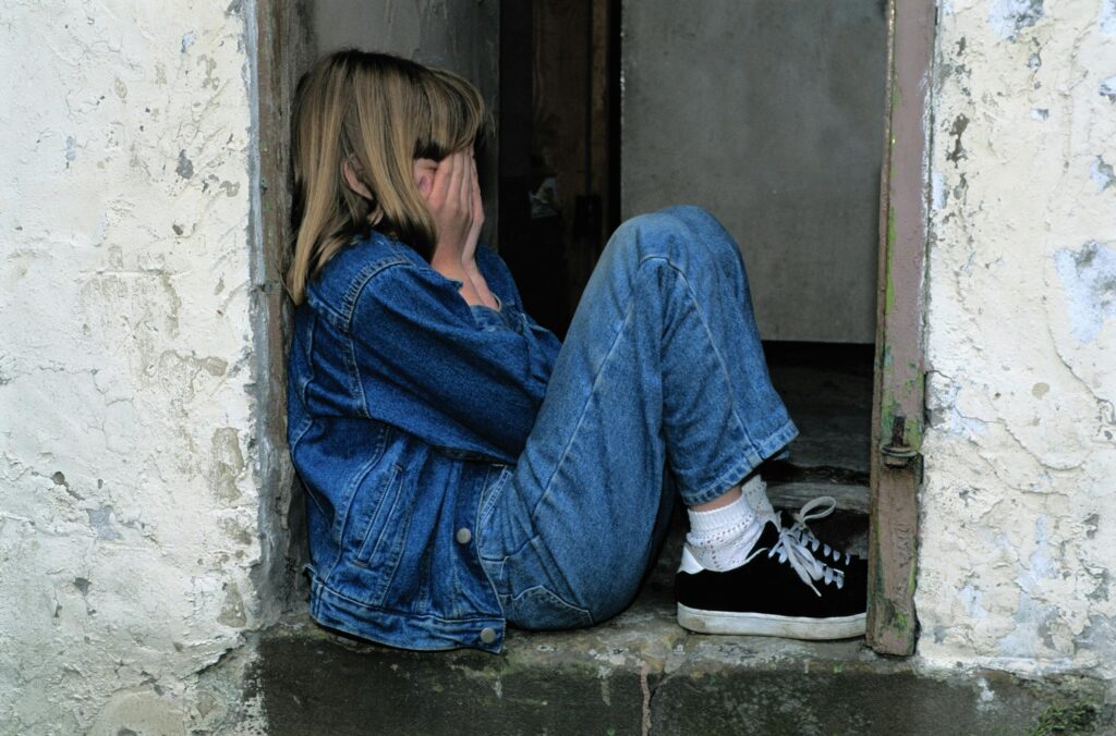 A 10-year-old girl is sitting in a doorway with their knees to their chest and their hands covering their eyes.